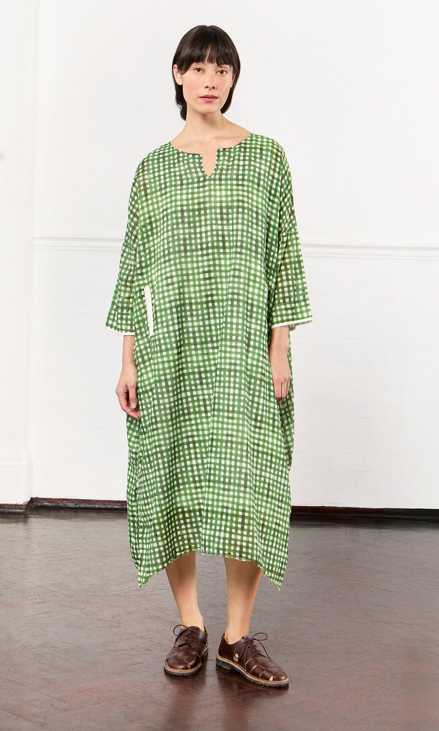 Plümo- Person wearing a loose-fitting Green Gingham Dress with brown shoes, standing indoors against a white wall.