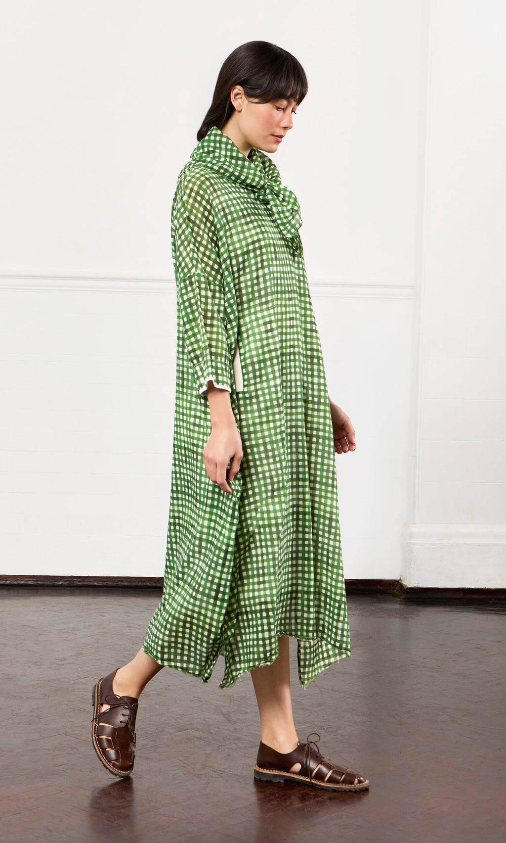 Plümo- Woman in a Green Gingham Dress and brown sandals walks indoors against a white wall.