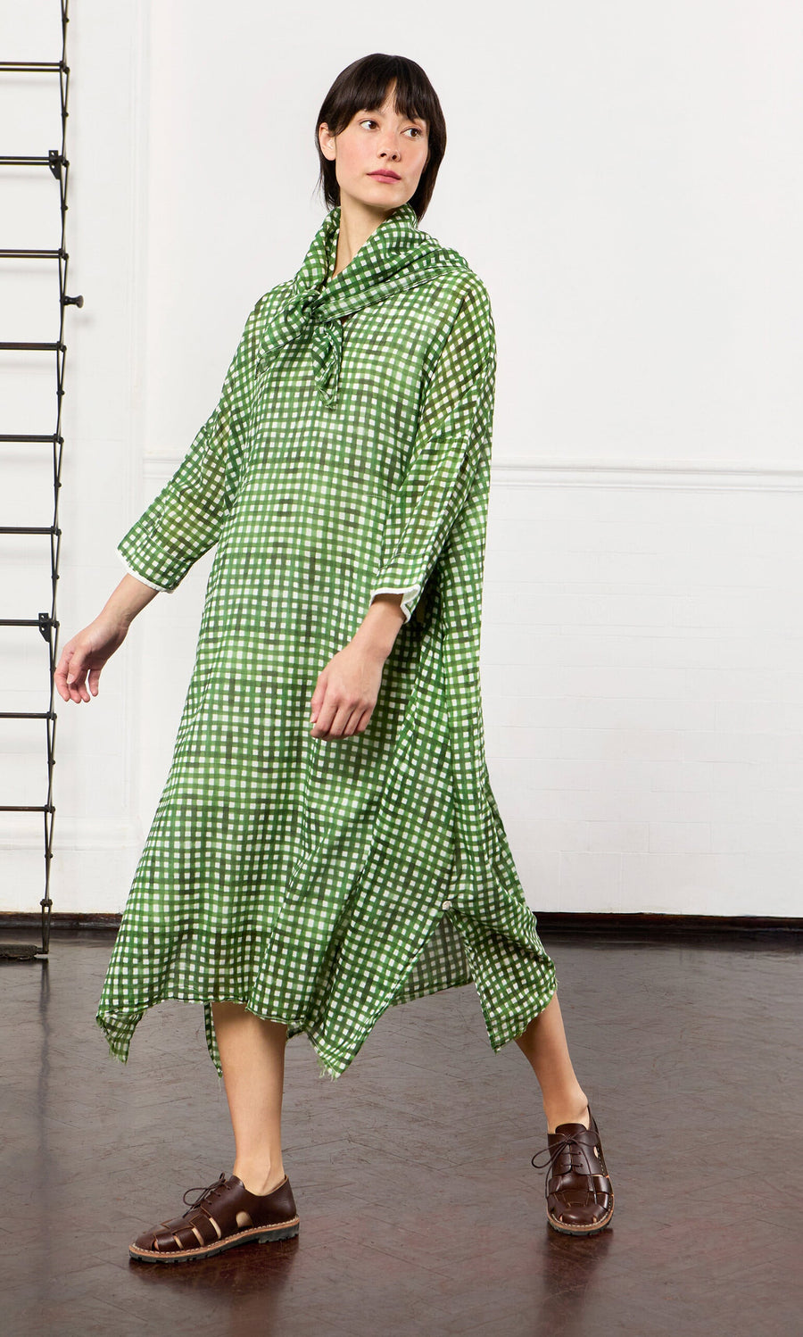 Plümo- Woman wearing a Green Gingham Dress and brown shoes, walking indoors on a dark wood floor.