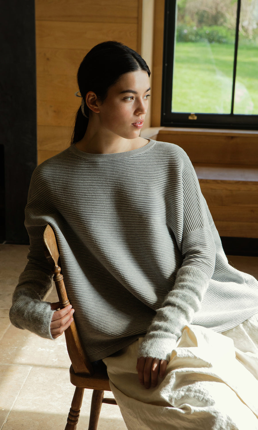 Plümo  A woman in a loose gray sweater and Grey Wrist Warmers sits by a window, gazing thoughtfully to the side.