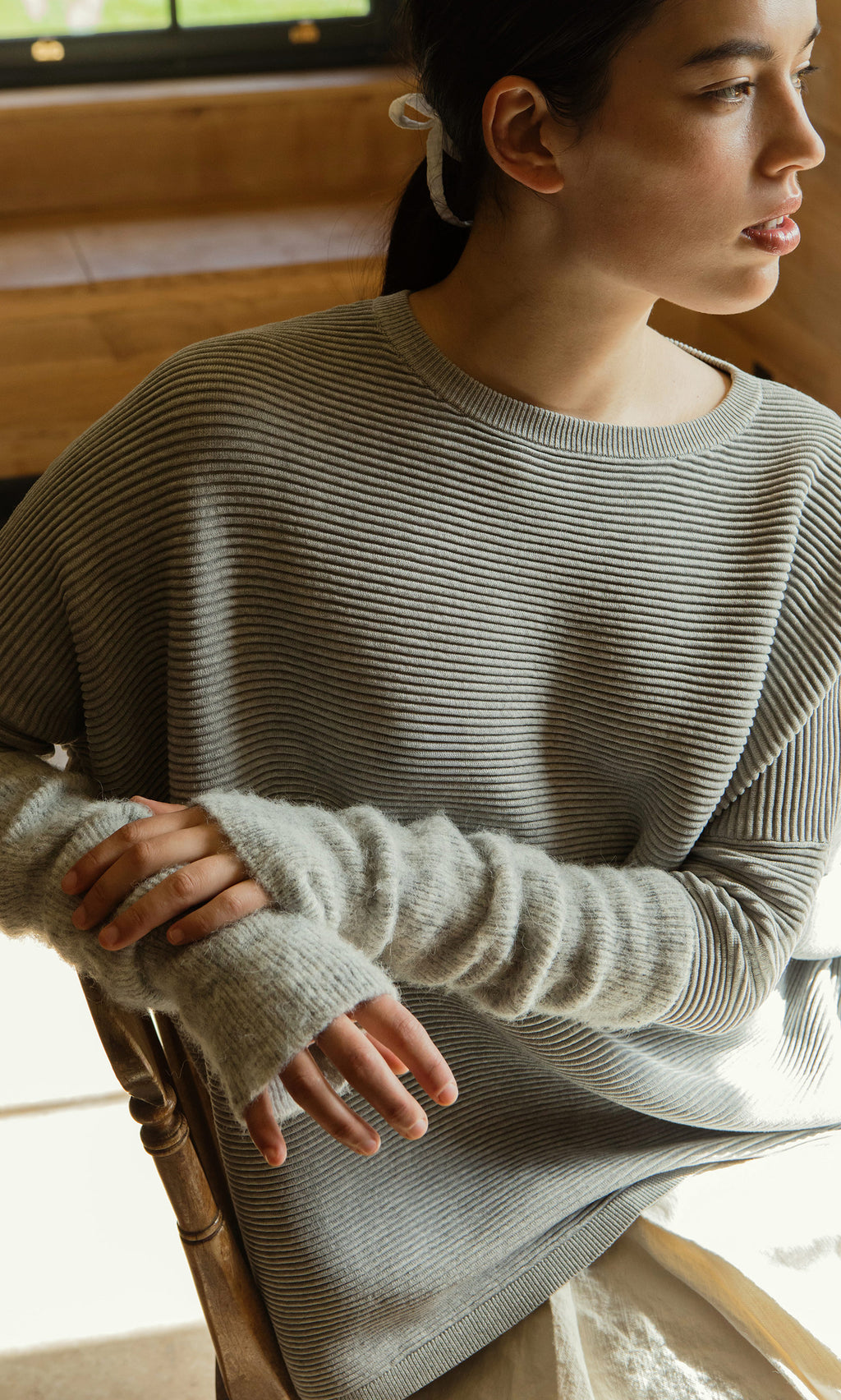 Plümo  Woman in a striped sweater and Grey Wrist Warmers sits sideways, sunlight highlighting her hands and face.