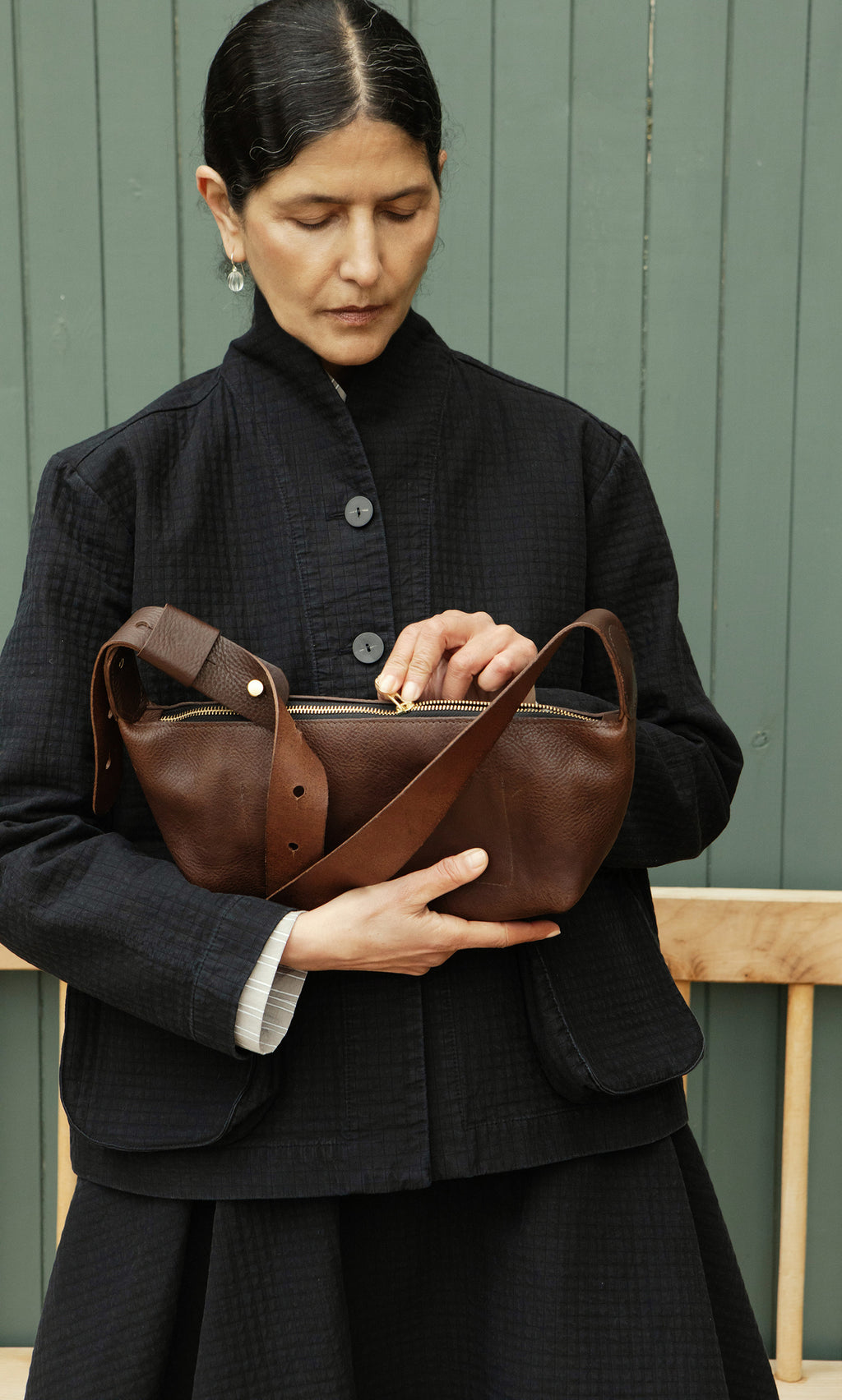 Plümo  A person in dark clothing opens a Hattie Bag by a wooden bench and green wall.