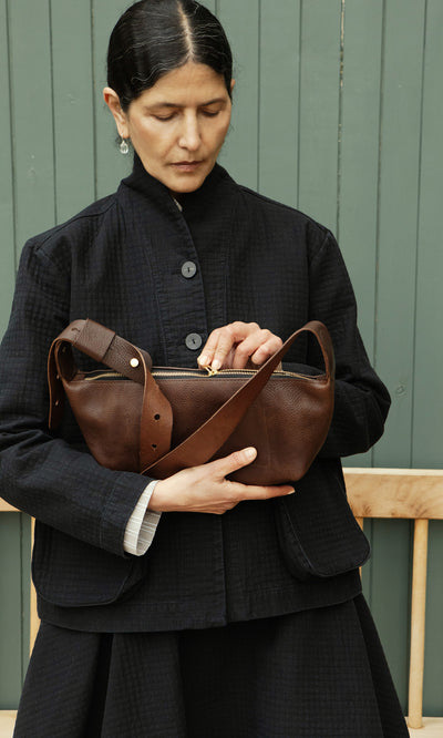 Plümo  A person in dark clothing opens a Hattie Bag by a wooden bench and green wall.