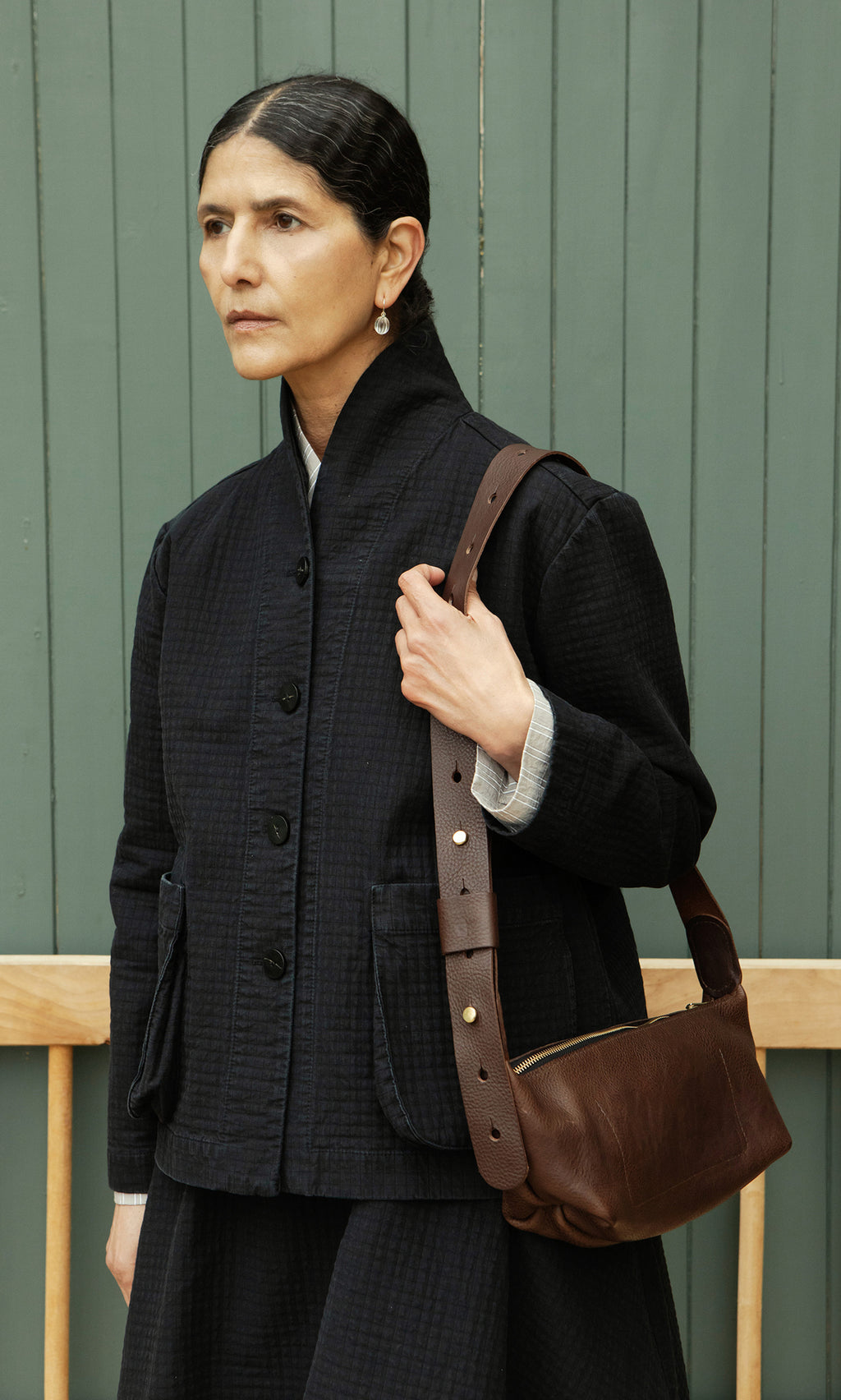 Plümo  Woman with dark hair in a bun holds the Hattie Bag, an Italian vegetable-tanned leather crossbody, by a green wall.