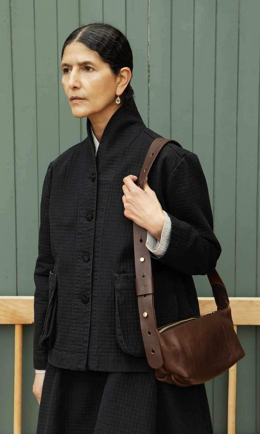 Plümo  Woman with dark hair in a bun holds the Hattie Bag, an Italian vegetable-tanned leather crossbody, by a green wall.
