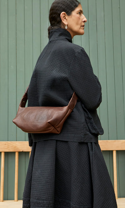 Plümo  Woman in black by a green wall wears the Hattie Bag, an Italian veg-tanned leather crossbody sling.
