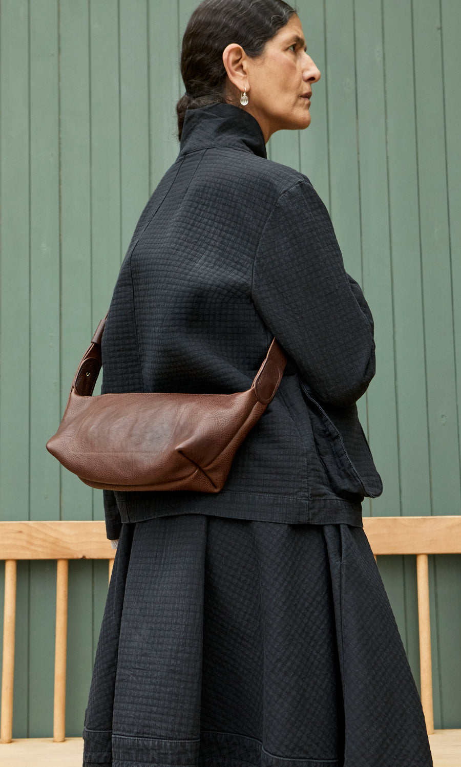 Plümo  Woman in black by a green wall wears the Hattie Bag, an Italian veg-tanned leather crossbody sling.