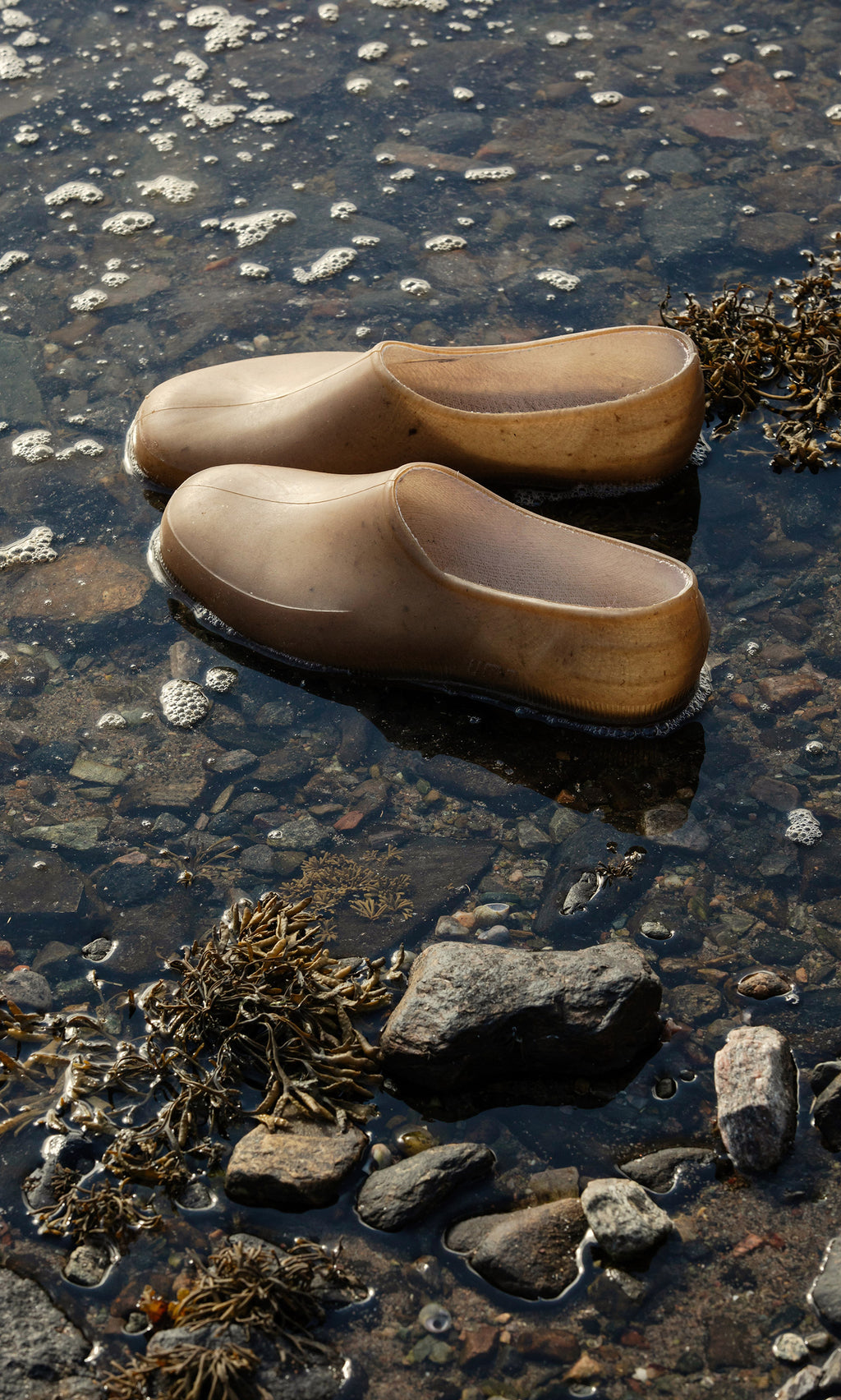 Plümo  A pair of brown Hemp Clogs sit in shallow water among rocks and seaweed at the shoreline’s edge.
