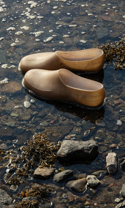Plümo  A pair of brown Hemp Clogs sit in shallow water among rocks and seaweed at the shoreline’s edge.