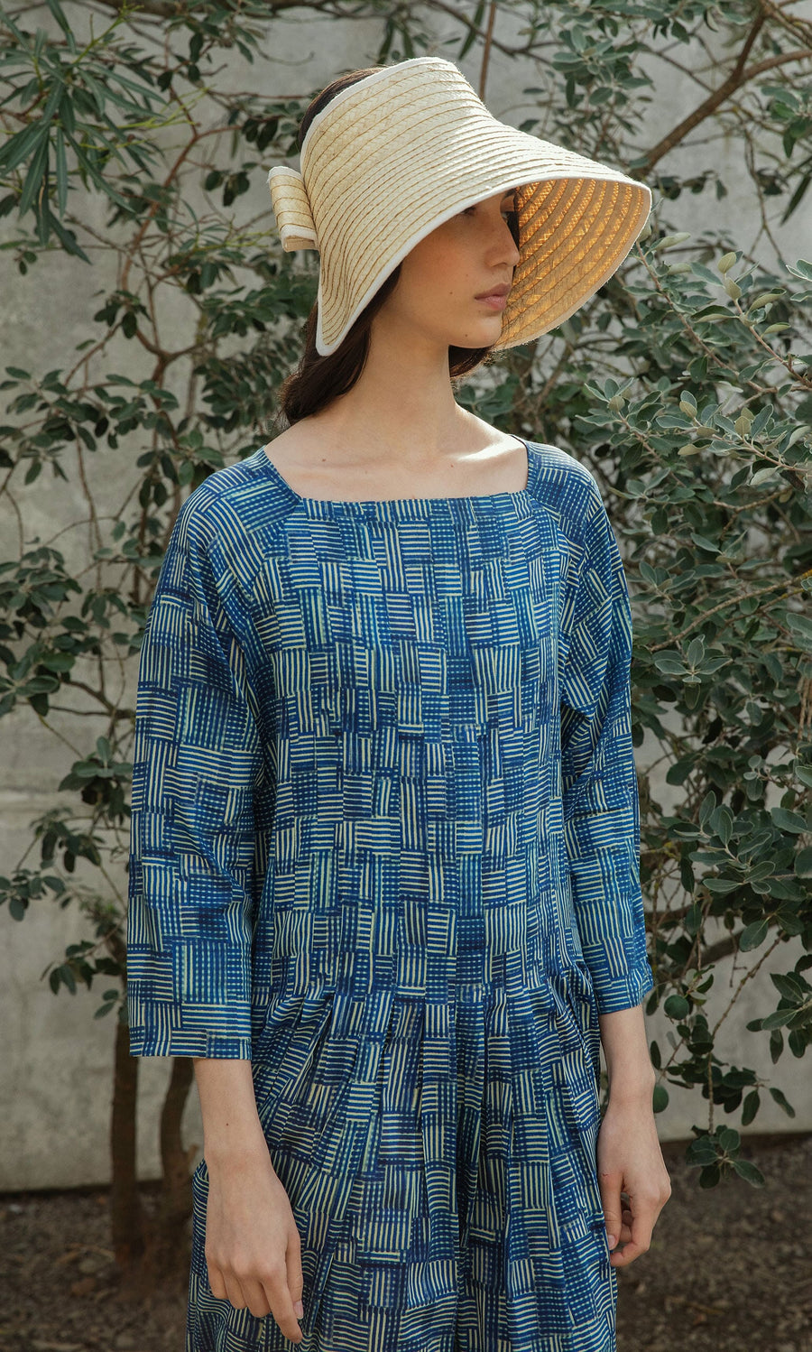 Plümo- Woman in a blue patterned dress wears the Bay Sun Hat, standing before lush greenery.