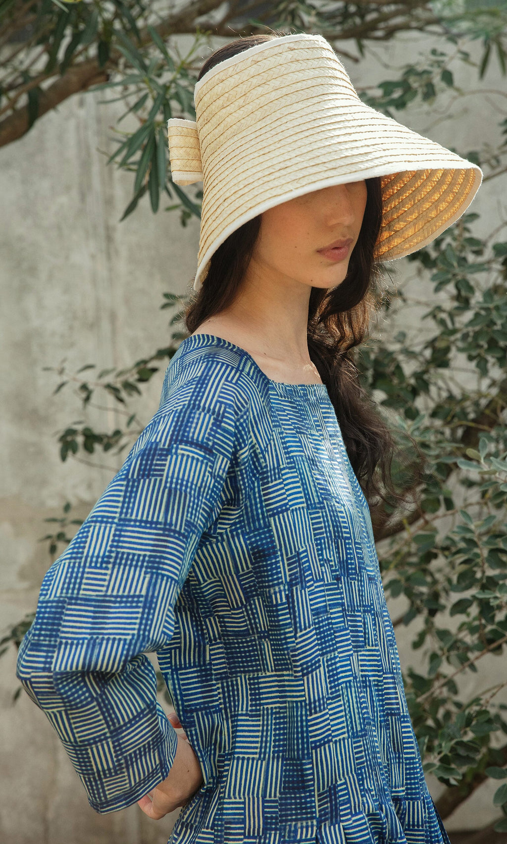 Plümo- Woman in a blue patterned dress wears the Bay Sun Hat while standing outdoors by green leafy plants.