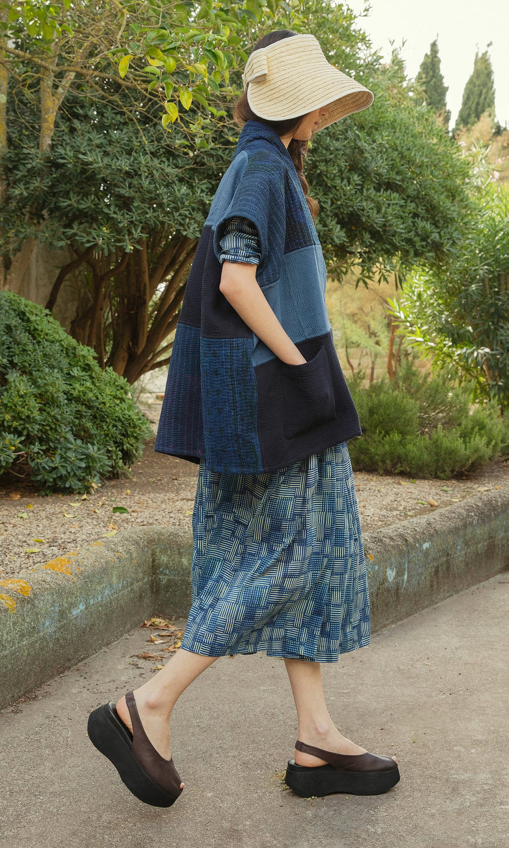 Plümo- A woman in a Bay Sun Hat, patchwork jacket, patterned skirt, and platform sandals walks outdoors.