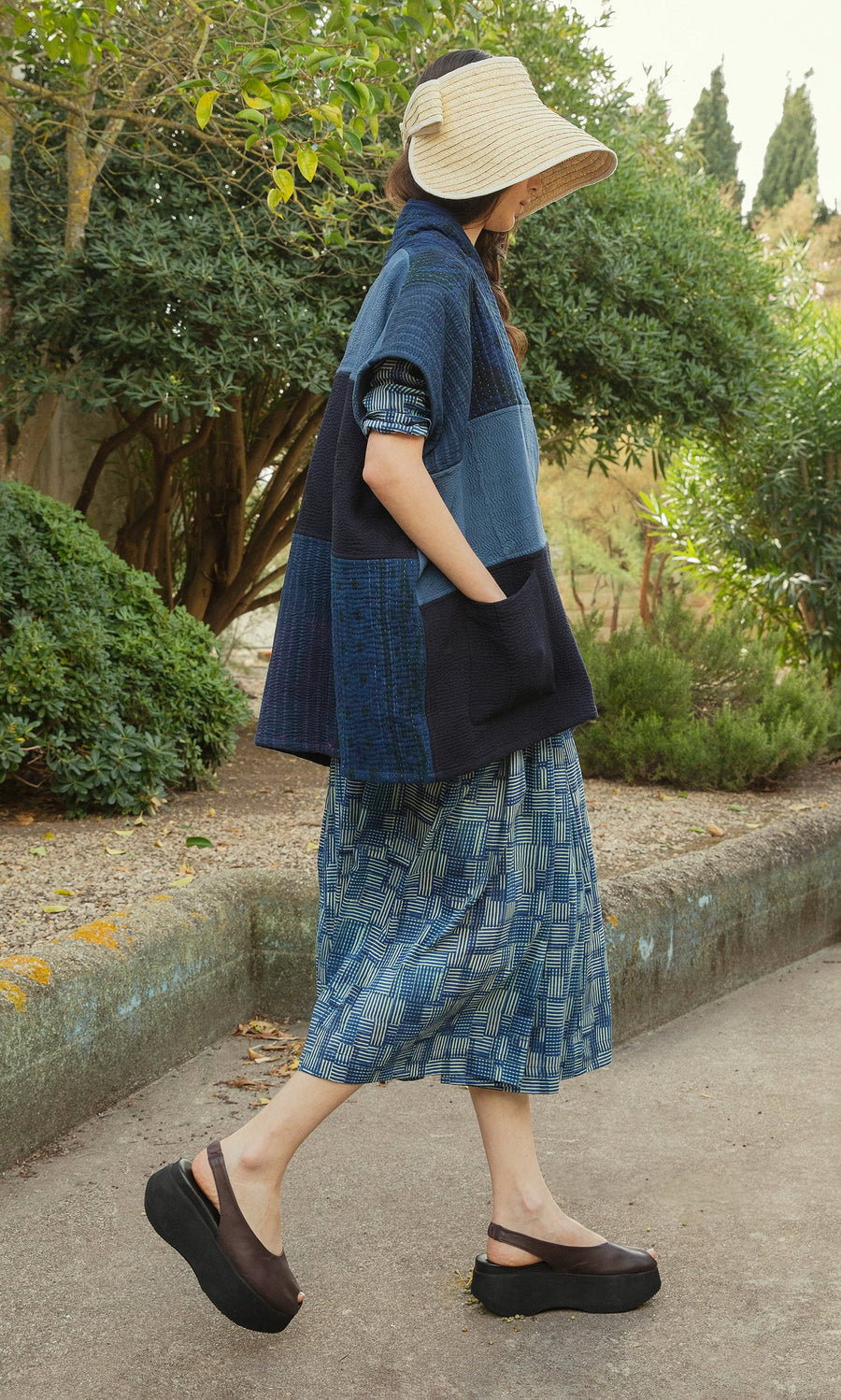 Plümo- A woman in a Bay Sun Hat, patchwork jacket, patterned skirt, and platform sandals walks outdoors.