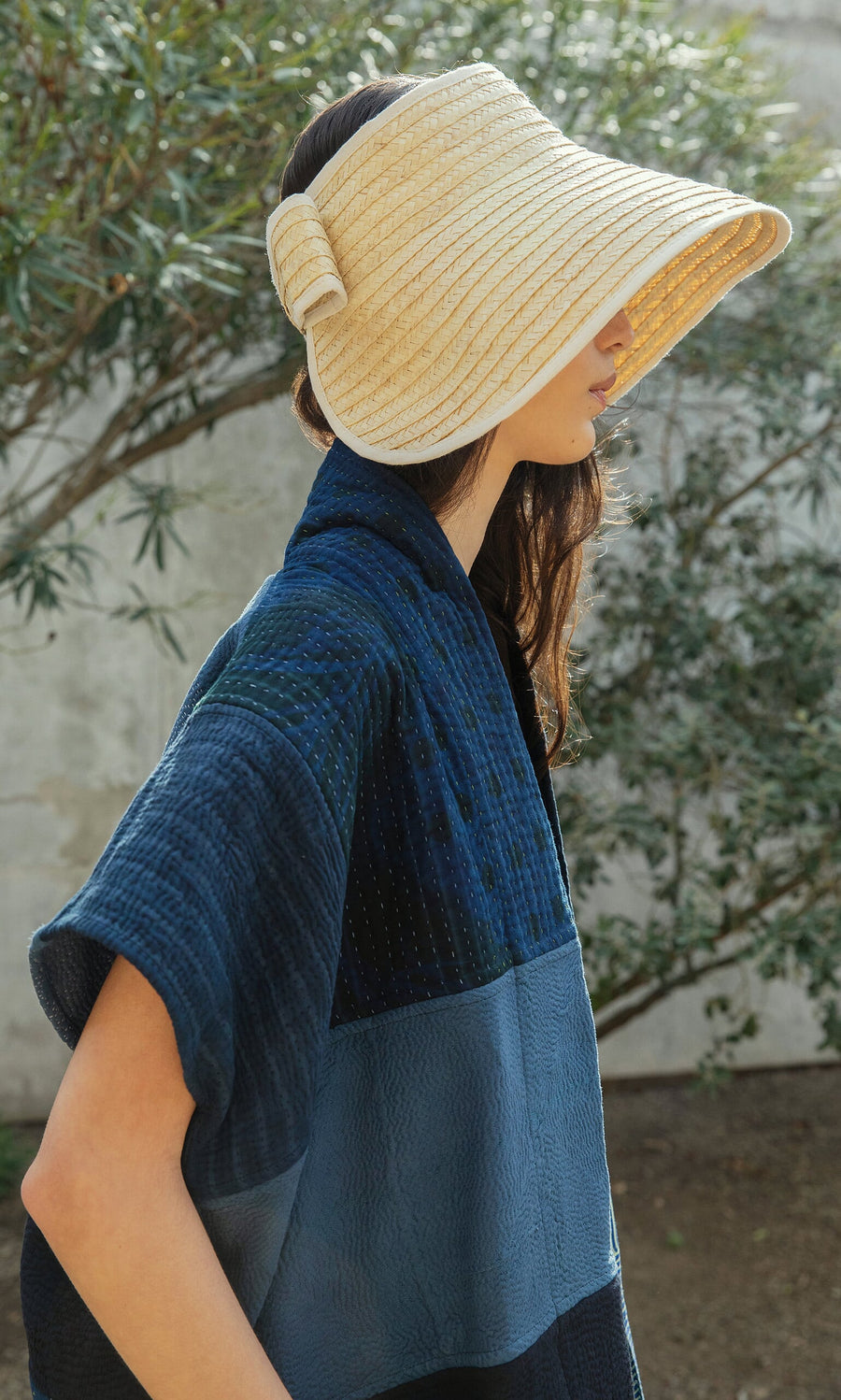 Plümo- Woman wearing the Bay Sun Hat stands outdoors near greenery, shown in profile with a blue textured jacket.