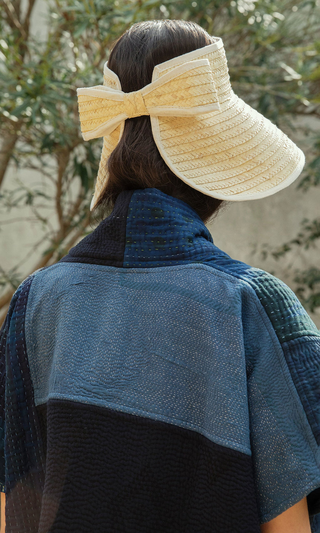 Plümo- Someone in a textured blue top wears the Bay Sun Hat, featuring an adjustable fit and a large bow, viewed from behind.