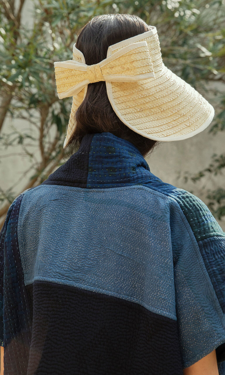 Plümo- Someone in a textured blue top wears the Bay Sun Hat, featuring an adjustable fit and a large bow, viewed from behind.