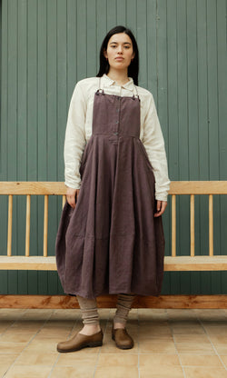 Plümo  A person in a white shirt and Ingham Dungaree Dress stands on tiled floor by a bench, wearing brown shoes and socks.