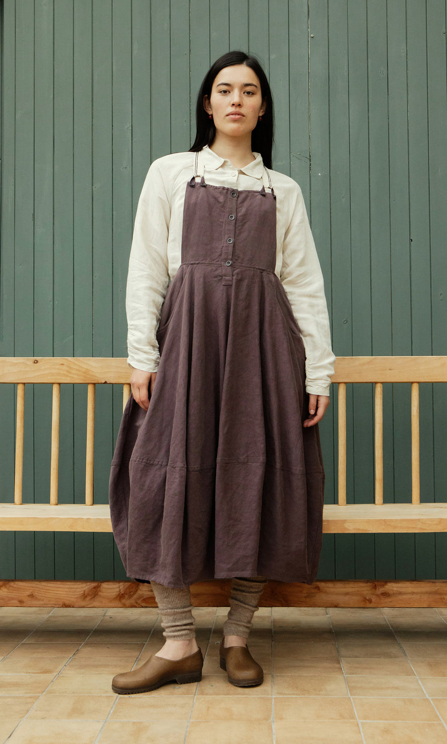Plümo  A person in a white shirt and Ingham Dungaree Dress stands on tiled floor by a bench, wearing brown shoes and socks.