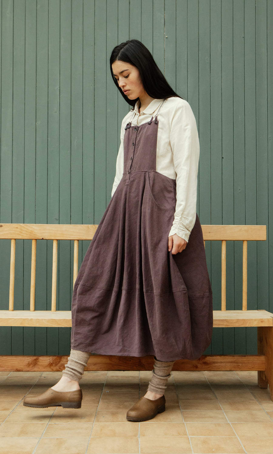 Plümo  Woman in a long brown dress, cream shirt, and Knitted Leg Warmers - Brown stands by a bench near a green paneled wall.