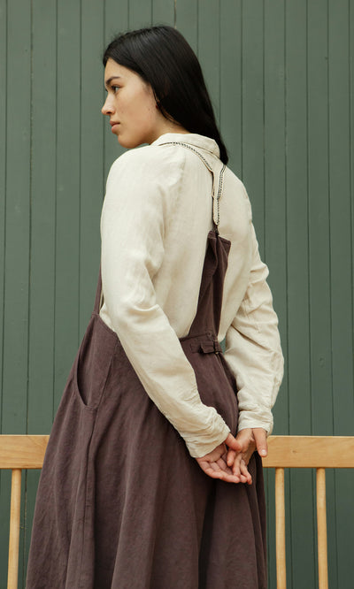Plümo  Woman in beige shirt and Ingham Dungaree Dress stands sideways, hands behind back, by a green wall.