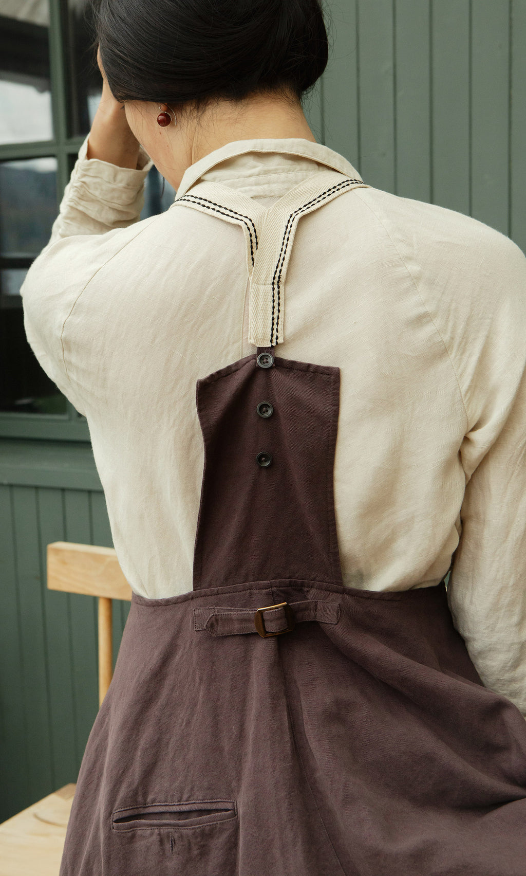 Plümo  Someone in a cream shirt and the Ingham Dungaree Dress sits facing away, adjusting their hair.