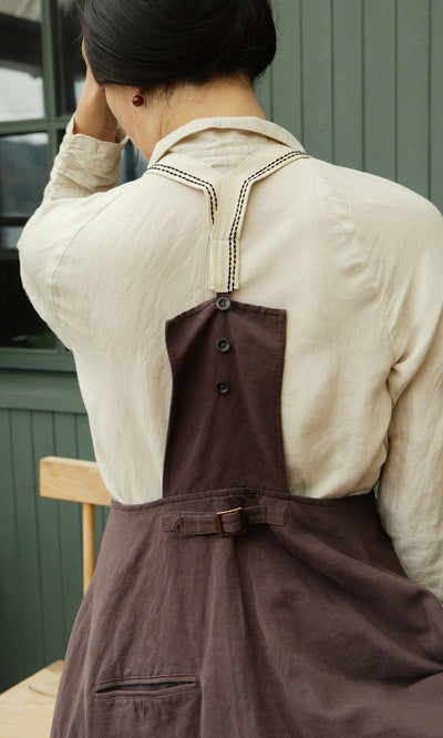 Plümo  Someone in a cream shirt and the Ingham Dungaree Dress sits facing away, adjusting their hair.