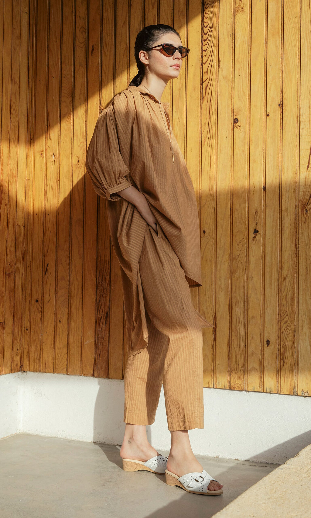 Plümo- Woman in sunglasses wears Caramel Pants and sandals, standing against a wooden wall in sunlight.