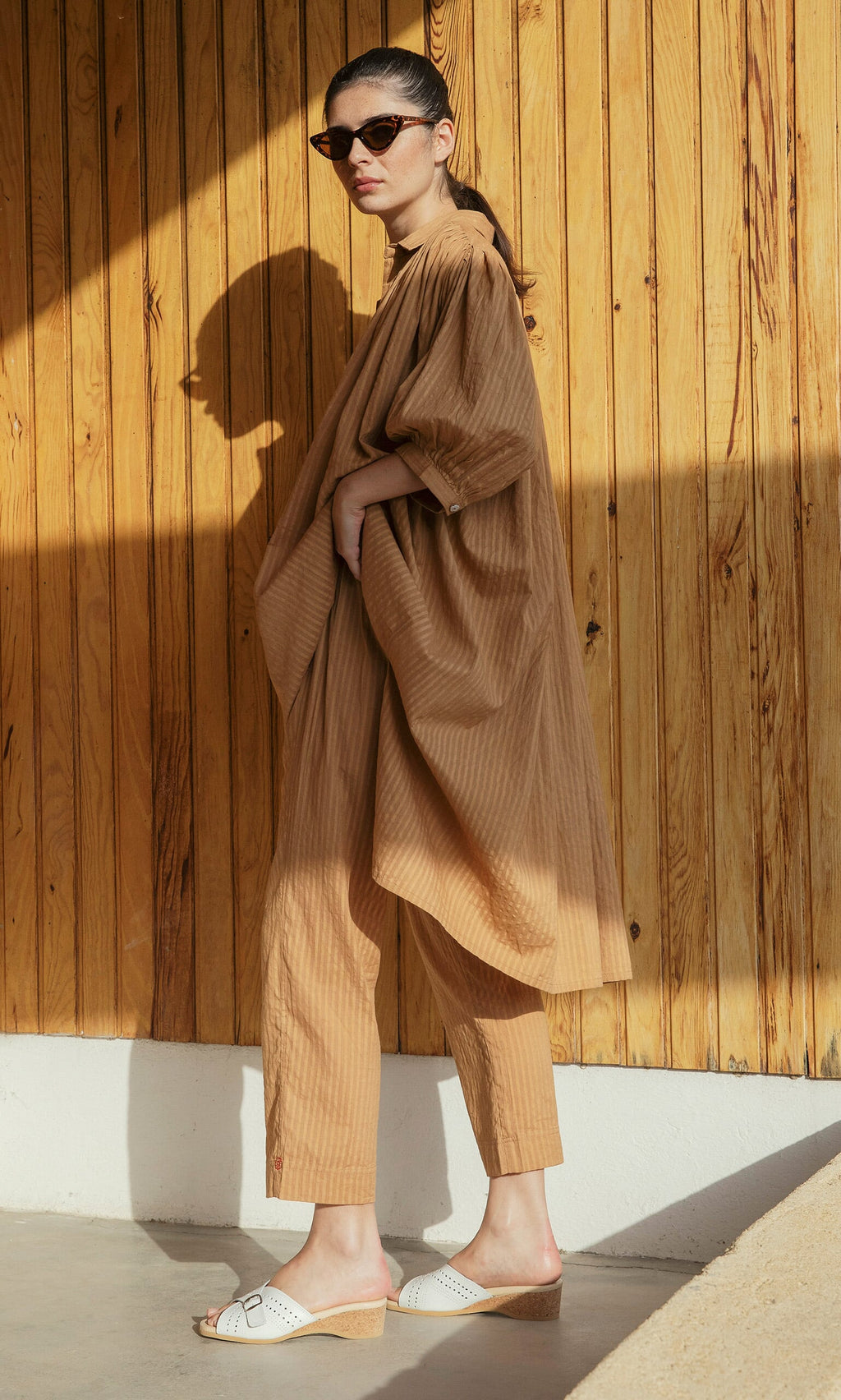 Plümo- Woman in sunglasses wears Caramel Pants and white sandals, standing by a wooden wall in sunlight.