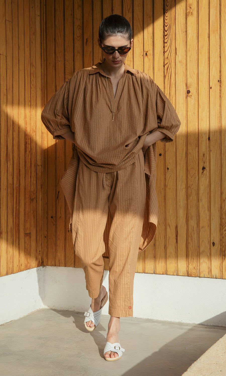 Plümo- Woman in Caramel Pants and white sandals, standing in sunlight with sunglasses against a wooden wall.