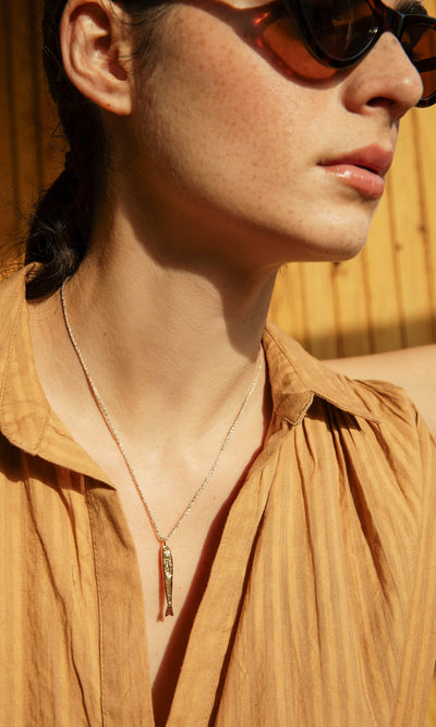 Plümo- Woman in sunglasses and tan shirt wears the Sardine Necklace, sunlight highlighting her face and neck.