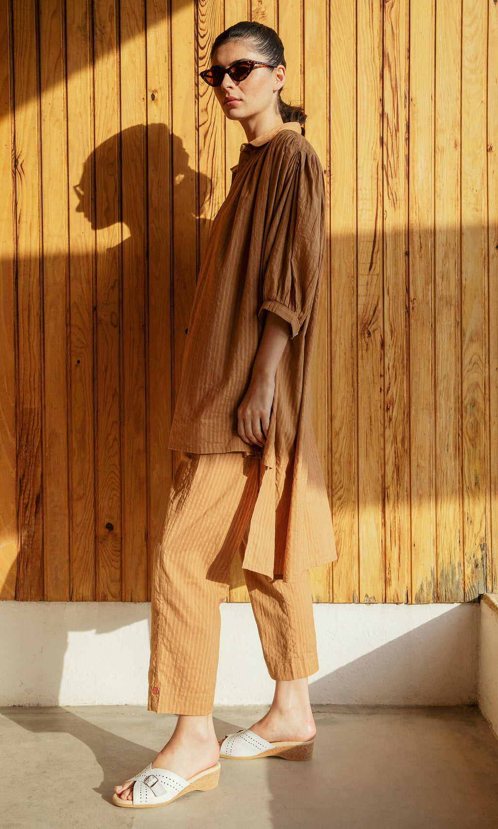Plümo- Woman in Caramel Pants, sunglasses, and white sandals stands against a wooden wall in sunlight.