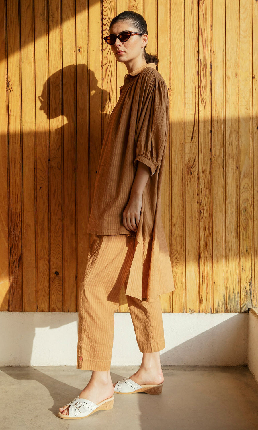 Plümo- Woman in Caramel Pants, sunglasses, and white sandals stands against a wooden wall in sunlight.