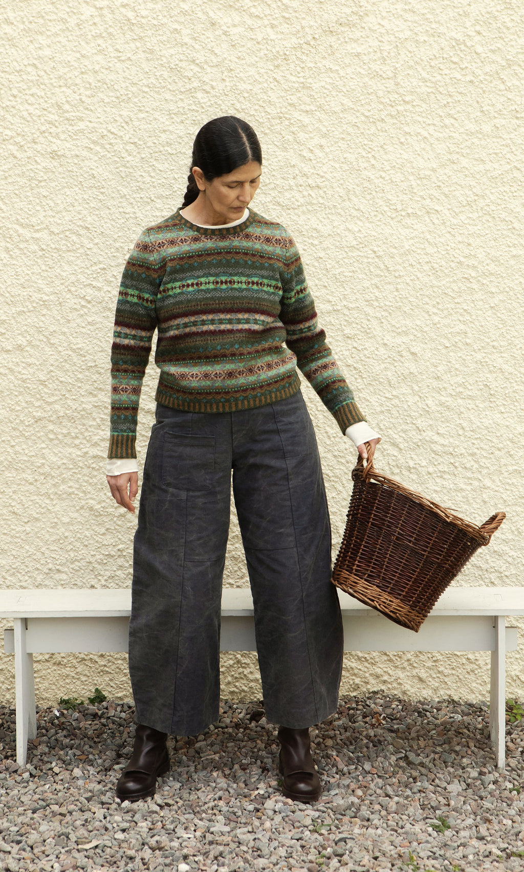 Plümo  Woman in an Iona Fairisle Jumper and wide-leg pants holds an empty wicker basket, standing on gravel by a white wall.