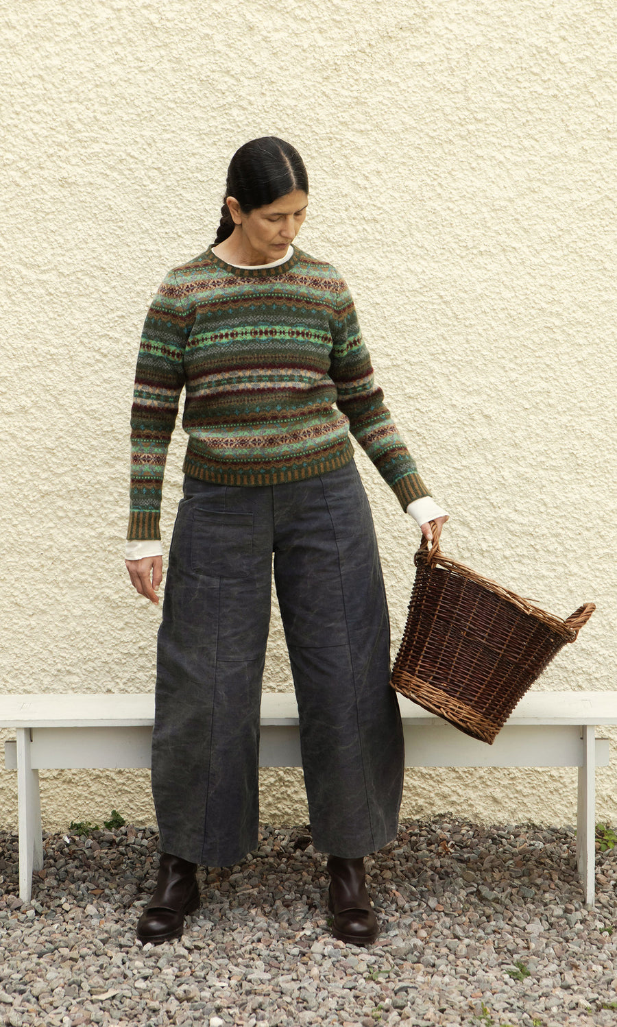 Plümo  Woman in an Iona Fairisle Jumper and wide-leg pants holds an empty wicker basket, standing on gravel by a white wall.