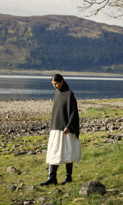 Plümo  A person in the Jack Rollneck - Graphite and a white skirt stands on grass by a lake with hills in the background.