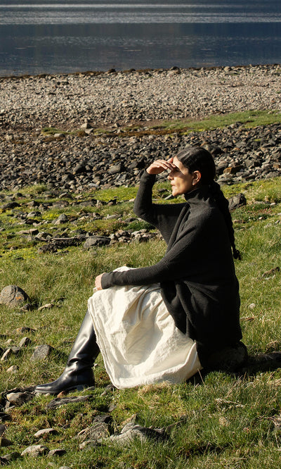 Plümo  Person in the Jack Rollneck - Graphite sits on grass by a rocky shore, shielding eyes and gazing at the water.
