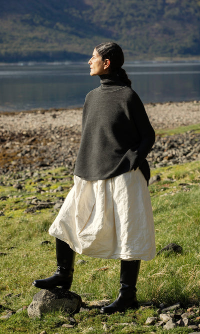 Plümo  A person in the Jack Rollneck - Graphite stands on grass near a rocky lakeshore, gazing into the distance.