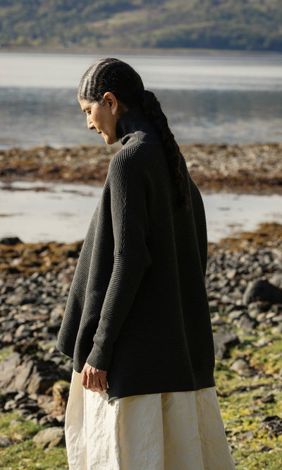 Plümo  Woman with braided hair stands by a lakeshore, wearing the Jack Rollneck - Graphite and a cream skirt, looking down.