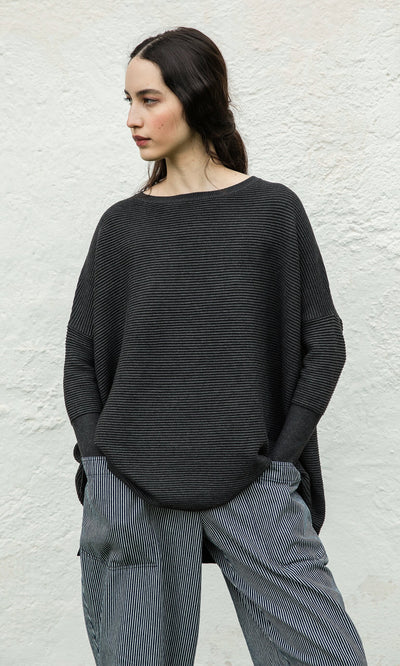 Plümo- Woman in the Jack Sweater - Graphite stands against a textured white wall, looking left.