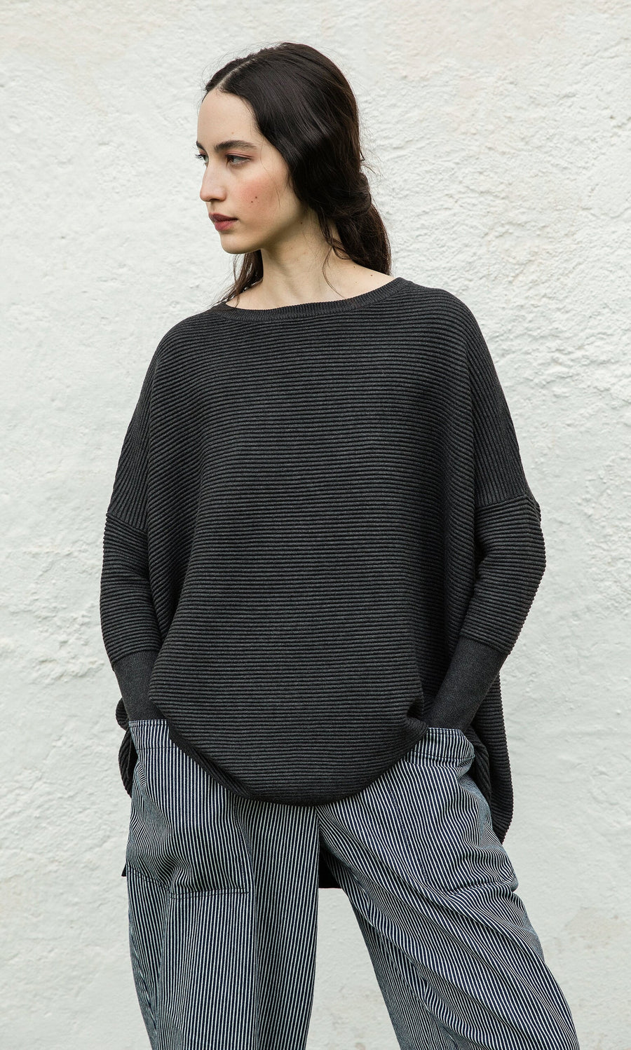 Plümo- Woman in the Jack Sweater - Graphite stands against a textured white wall, looking left.