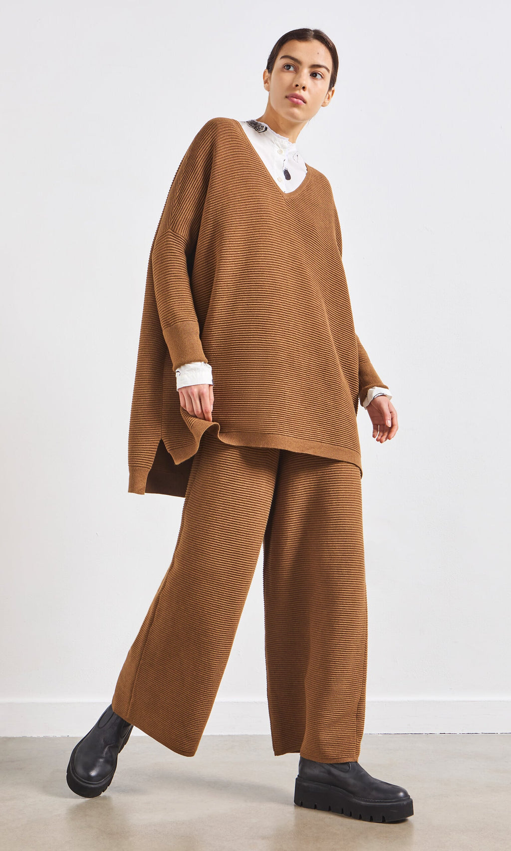 Plümo- A person in a loose brown sweater, Orla Pants - Chestnut, black boots, and a white collared shirt.