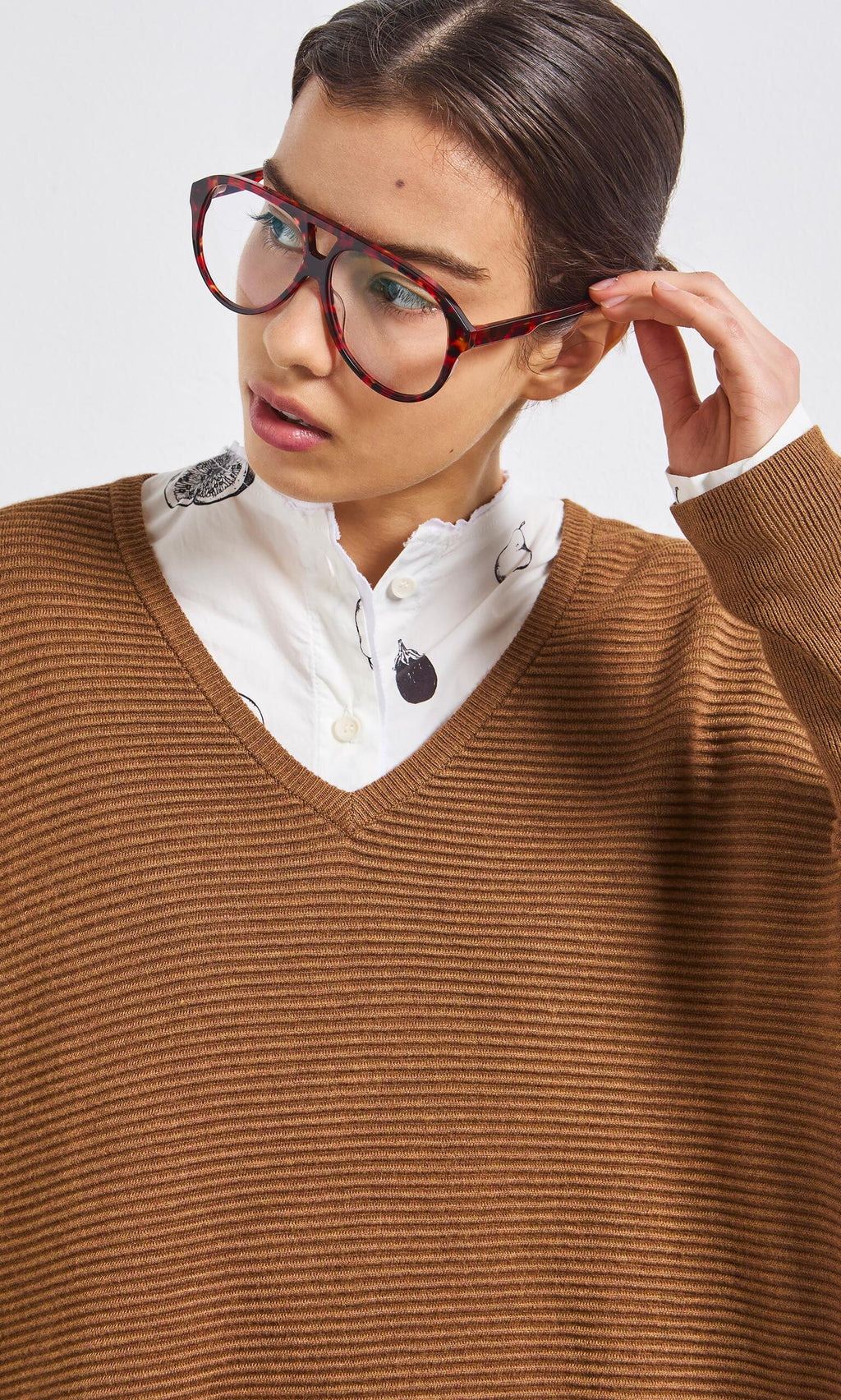 Plümo- Woman wearing Havana Red Readers, a white patterned shirt, and brown V-neck sweater, looking to the side.