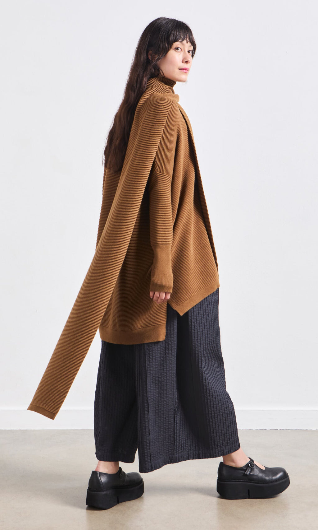 Plümo- Woman in a brown cardigan, Janie Scarf - Chestnut, wide black pants, and platform shoes.
