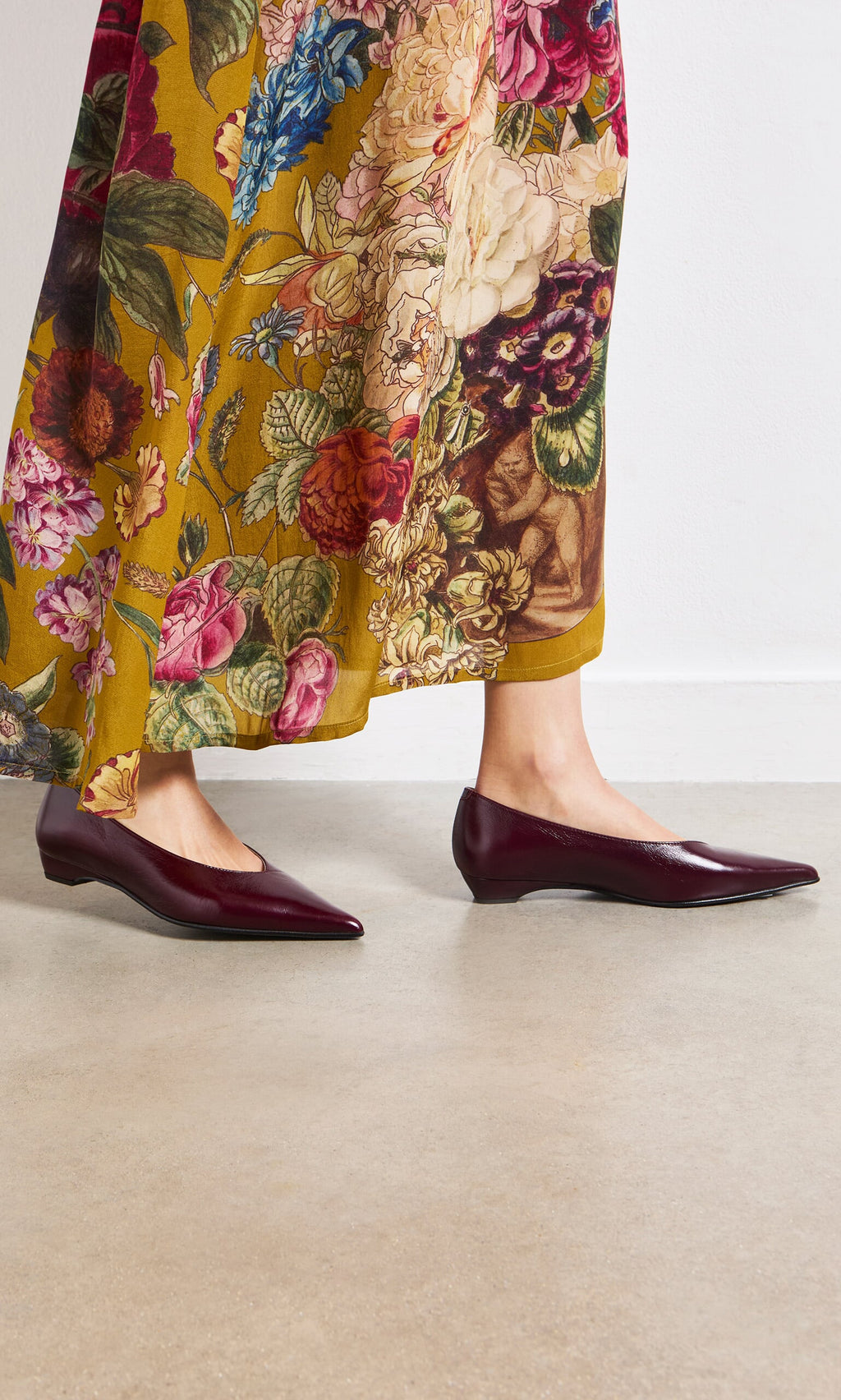 Plümo- Person wearing Jan Pumps in burgundy patent leather with a low block heel and a yellow floral skirt on a light floor.