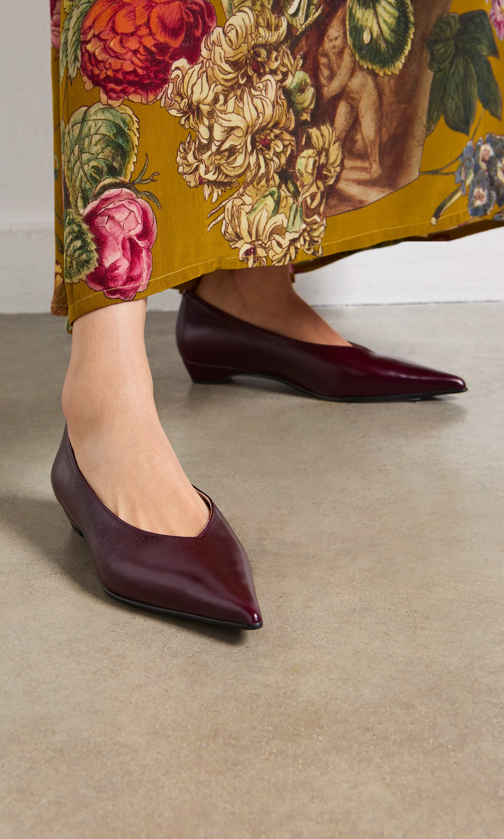 Plümo- Person wearing Jan Pumps in burgundy low block heels with a yellow floral skirt, standing on a beige floor.