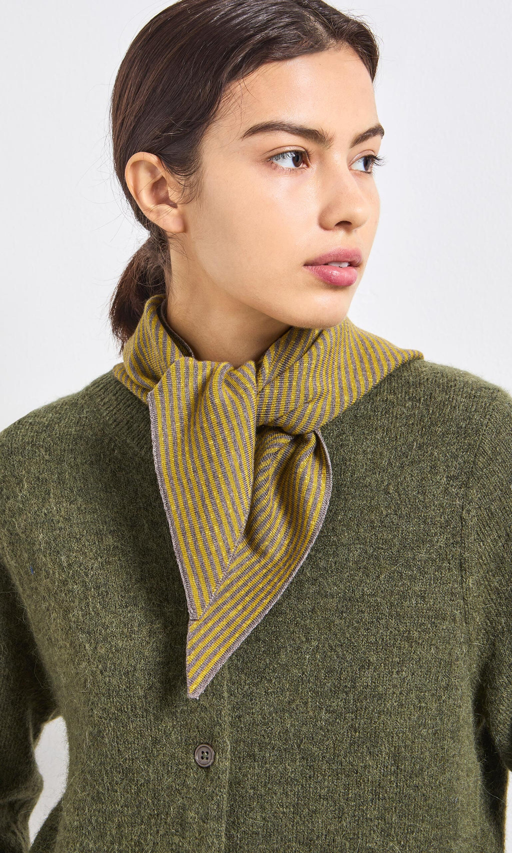 Plümo- A woman in an olive green cardigan and yellow-striped scarf, looking to the side.