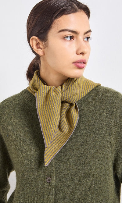 Plümo- A woman in an olive green cardigan and yellow-striped scarf, looking to the side.