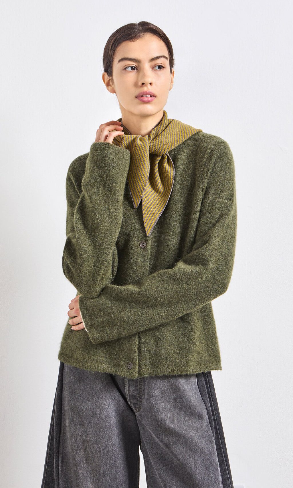 Plümo- Woman wears an olive green cardigan, gray jeans, and a yellow scarf against a white background.