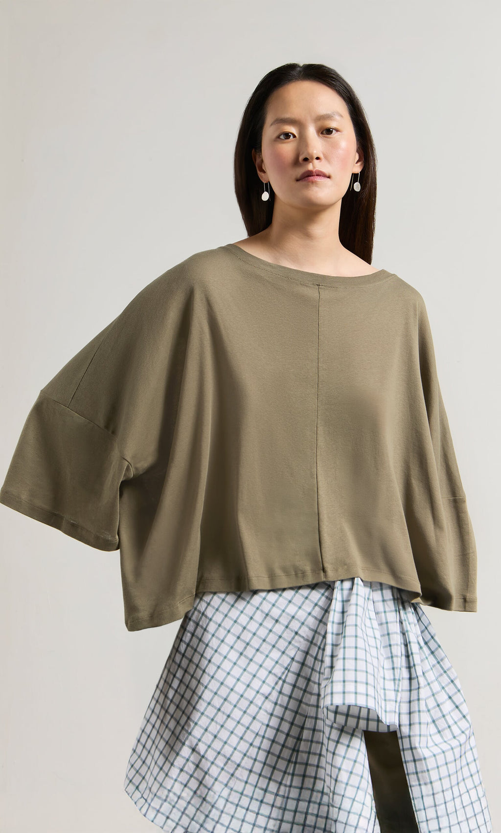 Plümo- Woman in a Kaia Jersey oversized boatneck top and blue plaid white skirt, standing against a plain background.