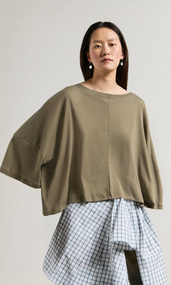 Plümo- Woman in a Kaia Jersey oversized boatneck top and blue plaid white skirt, standing against a plain background.
